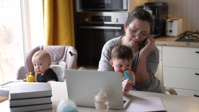 Busy mother multitasking with laptop and phone while holding toddler with spoon in modern kitchen. Working mom balances remote work and childcare duties as baby sits nearby in highchair.