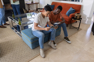 Obraz premium Diverse male friends sitting on blue ottoman in living room, sharing tablet and using notebook