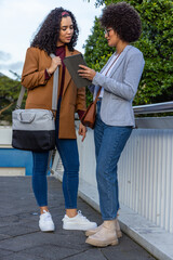 Fototapeta premium Diverse female colleagues leaning in, discussing tablet on walkway by metal railing wearing blazers