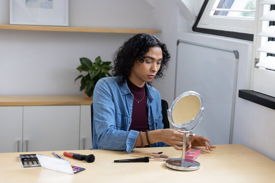 Non-binary person in denim at desk by window applying makeup with mirror, pink palette, brushes