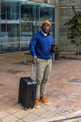 African American man waiting on brick plaza outside office in orange beanie holding suitcase phone © wavebreak3