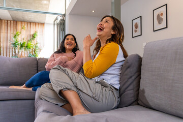 Obraz premium Diverse female friends laughing on gray sofa at home wearing mustard sleeves and pink sweater