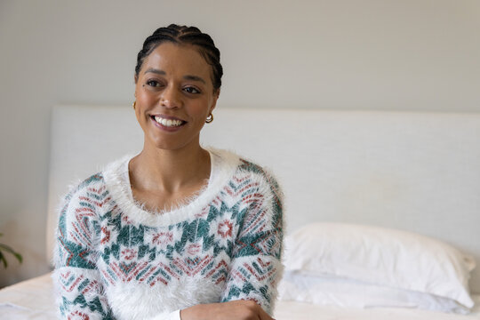 African American woman sitting and smiling on bed in muted bedroom, wearing fuzzy patterned sweater