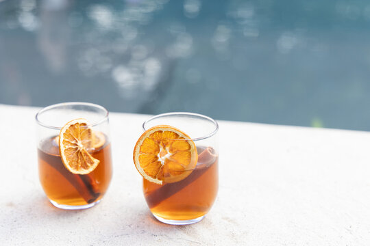 Two glasses sitting on pool ledge with amber drinks, orange wheels, cinnamon sticks, copy space