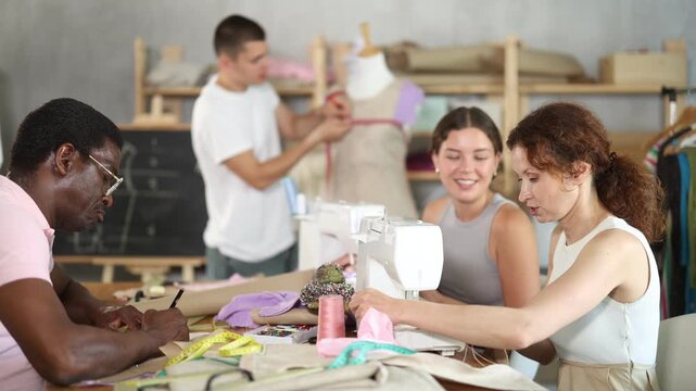 Positive interested female fashion design students stitching on machines and friendly talking, sharing sewing tips in hands-on workshop while male classmate adjusting garment on mannequin