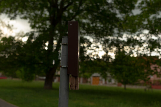 Close-up of a weathered wooden brush head attached to a metal post in a park setting at dusk. The small brush shows frayed bristles and worn wood grain
