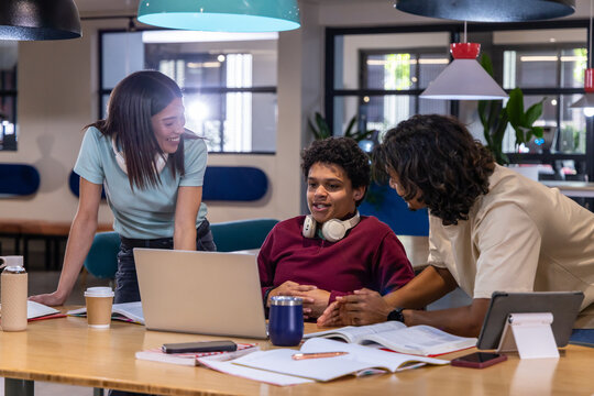 Diverse peers leaning over laptop, discussing notes at modern study lounge with headphones