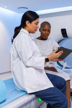 Asian Dr and African-American patient wearing gown reviewing tablet, sitting on bench near CT bore