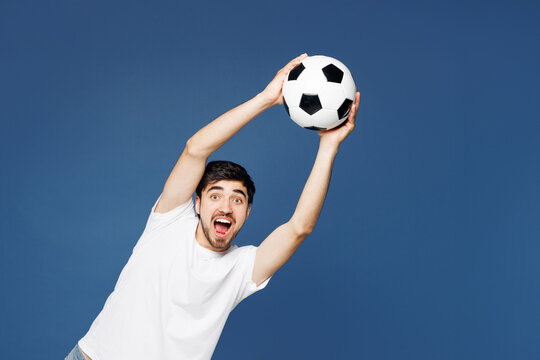 Young overjoyed cheerful smiling happy man fan wearing basic white t-shirt cheer up support football sport team hold in hand catch soccer ball watch tv live stream isolated on plain blue background