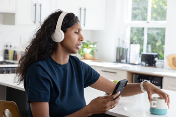 African American woman sitting in kitchen wearing navy tee, headphones, holding phone, reaching mug © wavebreak3