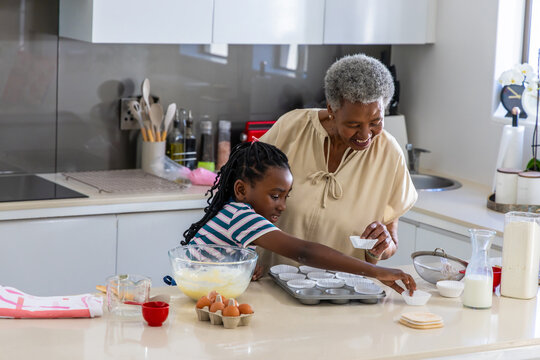 African American senior woman guiding teenage girl placing cupcake liners at kitchen countertop