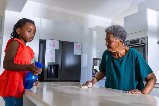 African grandma and granddaughter cleaning kitchen counter with blue sprayer and cloth in teal, red