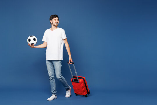 Traveler man wears white clothes cheer up support football sport team hold suitcase bag soccer ball isolated on plain blue background. Tourist travel abroad in free time rest Air flight trip concept.