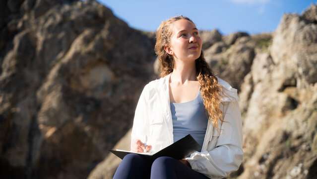 Portrait of a young female traveler taking notes on a notebook using a pen outdoors in a rocky mountainous environment.