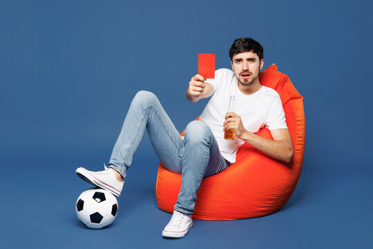 Full body sad upset young man fan wear white t-shirt cheer up football sport team hold in hand red card soccer ball watch tv live stream sit in bag chair drink beer isolated on plain blue background.