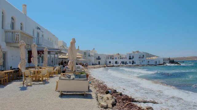Explore the beautiful coastline of Naousa, Paros, Greece, where charming cafes line the shore. Enjoy the vibrant blue waters as gentle waves lap against the sandy beach on a sunny day.