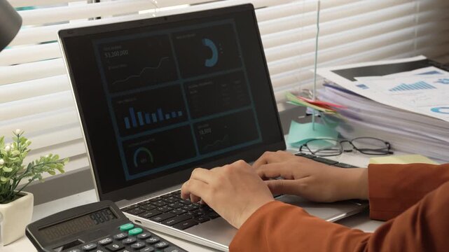 Close view of hands typing on a laptop with business charts on screen, calculator, papers, and office desk setup in soft daylight.
