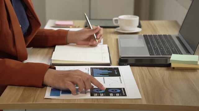 Businesswoman works at a desk with laptop, notebook and printed charts, writing notes during data analysis in a modern office setting.