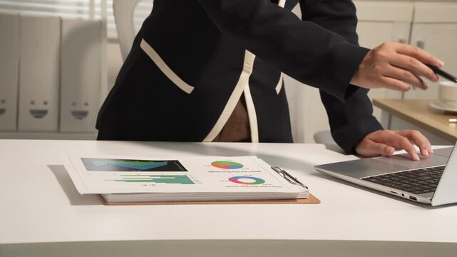 Businesswoman pointing at a laptop in a clean office with printed charts on the desk, showing data review, planning, teamwork and professional workplace activity