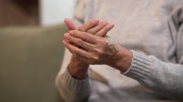 Close-up of an older person's clasped hands while sitting indoors in a soft sweater, showing age, calm, reflection, care, and quiet support in a home setting.