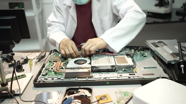 Technician in a lab coat and mask repairs an open desktop computer on a workbench, surrounded by parts and tools in a clean electronics lab.