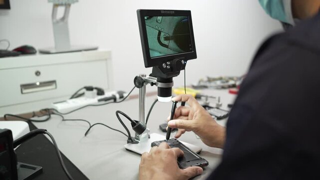 Technician in safety glasses and mask repairs a smartphone at a workbench using a microscope screen and tools in an electronics workshop.
