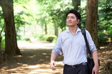 Fototapeta premium 緑豊かな公園を歩く30代の日本人男性会社員が鞄を肩にかけて爽やかな表情で遠くを見つめる様子