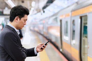 駅のプラットホームで真剣にスマートフォンを操作するスーツ姿で通勤中の30代の日本人の会社員の男性 © Hiroba Studio