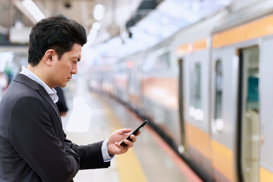 駅のプラットホームで真剣にスマートフォンを操作するスーツ姿で通勤中の30代の日本人の会社員の男性