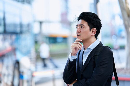 都市のバス停でバスを待ちながら考え事をする30代の日本人の男性の会社員