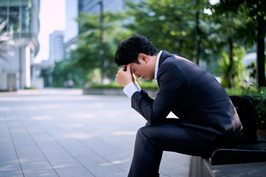 都会の屋外のベンチで仕事のストレスで頭を抱えて悩む20代の日本の男性の会社員