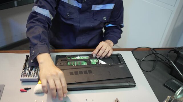Technician repairs a laptop at a workbench using a screwdriver, with tools and electronic parts spread across the table in a clean workshop
