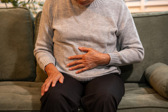 Senior woman sitting on a sofa and holding her stomach, showing abdominal pain, indigestion, cramps or discomfort at home in a close cropped view
