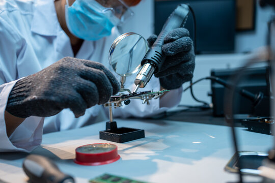 Technician in a lab coat and mask repairs a green circuit board with a soldering tool at a workbench, showing electronics service, maintenance, and precision work.