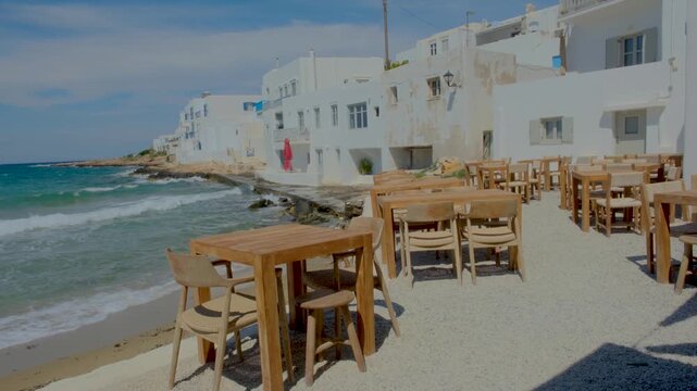 Relax at seaside tables in Naousa Paros Greece, where the sun shines on white buildings and gentle waves kiss the shore. A perfect spot for vacationers to enjoy fresh local cuisine and stunning views.