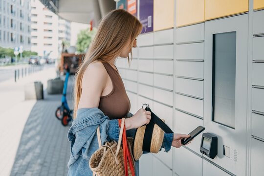 Woman presents smartphone to automated parcel station scanner while holding bags and straw hat. Click and collect retail service, online shopping parcel pickup, consumer convenience economy lifestyle.