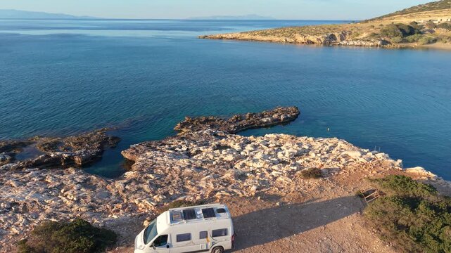 Breathtaking views of the tranquil waters and rocky coastline of Paros Island are showcased as a camper van rests at a serene spot. Experience the peaceful charm of the Cyclades during sunny hours.