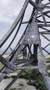 Croce metallica posizionata sulla cima di un pendio erboso, ripresa con movimento lento che mostra la struttura contro il cielo nuvoloso e la vista ampia sulla valle sottostante