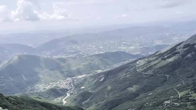 Vista dalla cresta di una montagna, con la camera che mostra pendii verdi, strade serpeggianti e valli profonde che si estendono verso rilievi lontani sotto un cielo parzialmente nuvoloso