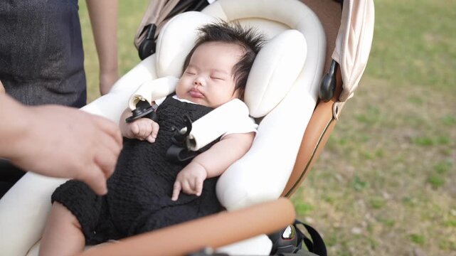 Calm baby sleeps in soft white car seat. Parent gently adjusts baby's clothing nearby. Baby's tiny hand rests near face. Peaceful moment outdoors, perfect for family scenes