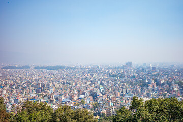Naklejka premium A wide panoramic view of Kathmandu, Nepal, stretching across the Kathmandu Valley beneath a soft blue sky. Captured from an elevated hillside viewpoint with green treetops in the foreground