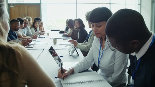 Diverse group of professionals actively engaged in a conference or workshop, taking notes and using technology. Focused individuals collaborate and learn together in a productive business environment.