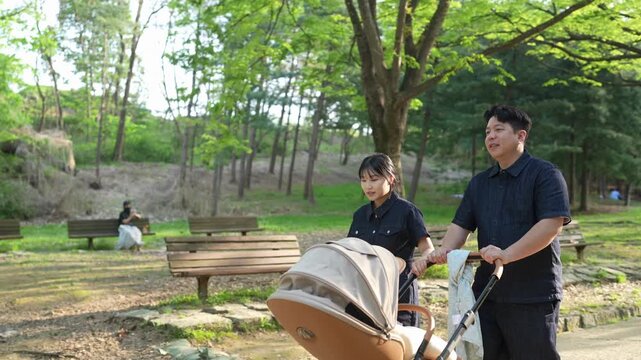 Parents stroll through green park with baby stroller. Sunlight filters through tall trees overhead. Woman smiles gently beside her husband. Man looks ahead, relaxed and content