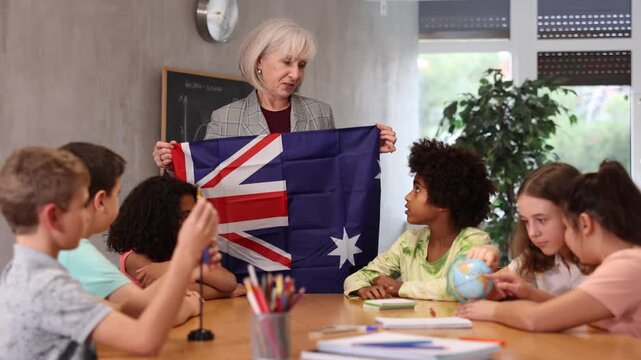 Elderly high teacher, conducting a lesson in the classroom, holds the national flag of Australia and tells the pupils the history of the country. High quality 4k footage