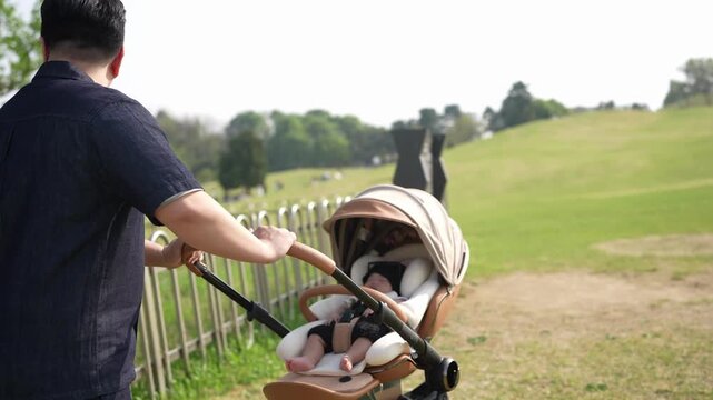 Man pushes stroller through park. Child sleeps in cozy carriage. Sunlight glows on green grass. Dad looks thoughtful, calm. Ideal for parenting or family scenes. Nature and peace evoke gentle mood