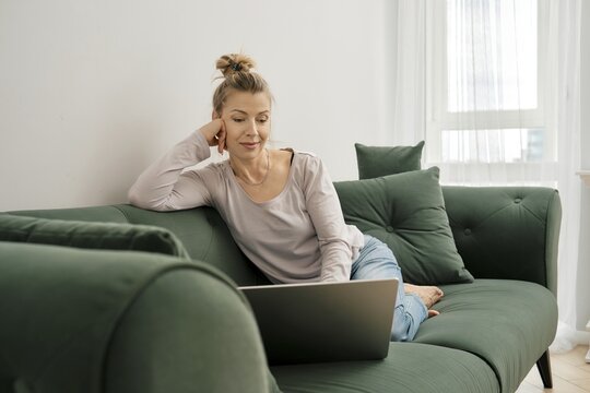 Middle-aged woman dressed casually sits on a green sofa, engaged with her laptop. Sunlight streams through a window, creating a cozy atmosphere in her living room