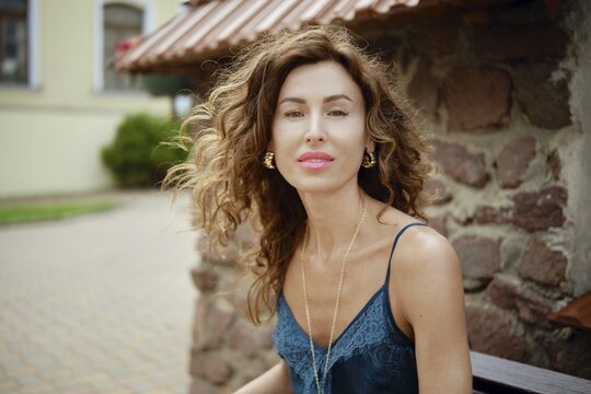 A woman with beautiful curly hair sits on a bench next to a rustic stone wall. She wears a stylish outfit and smiles warmly at the camera