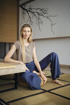 A woman is sitting comfortably on a floor mat in a modern room. She is dressed in casual clothes and has long hair. The room has minimal decor and natural light coming through the window
