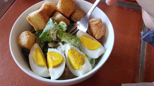Hand using fork stirring Caesar salad with lettuce, egg, crouton, parmesan and chicken grilled in the white bowl isolated on wooden table background