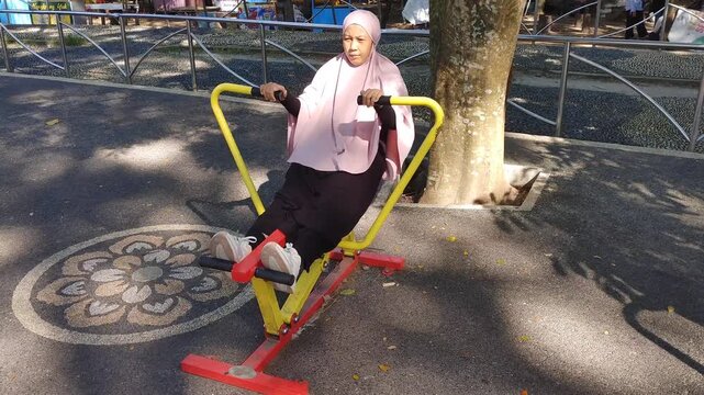 Happy active woman wearing large hijab exercising at outdoor fitness park on rowing machine. Healthy lifestyle concept